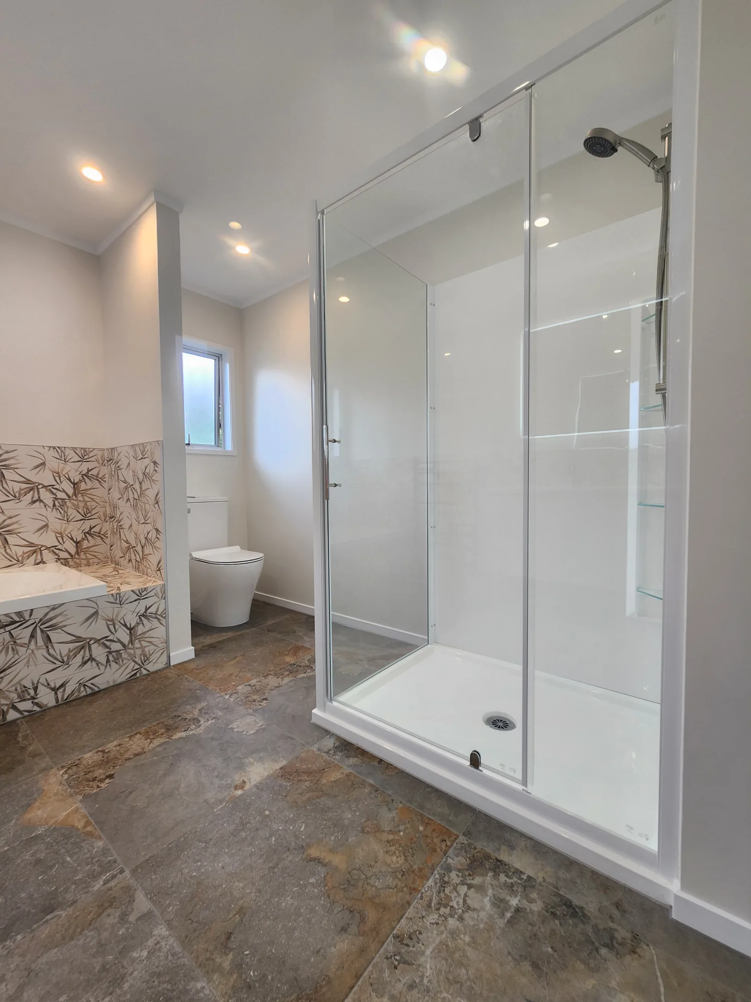 Walk-in shower with glass screen beside the botanical feature-tile bath surround and stone-look porcelain floor in a Massey bathroom, West Auckland — by Tarara Tiling