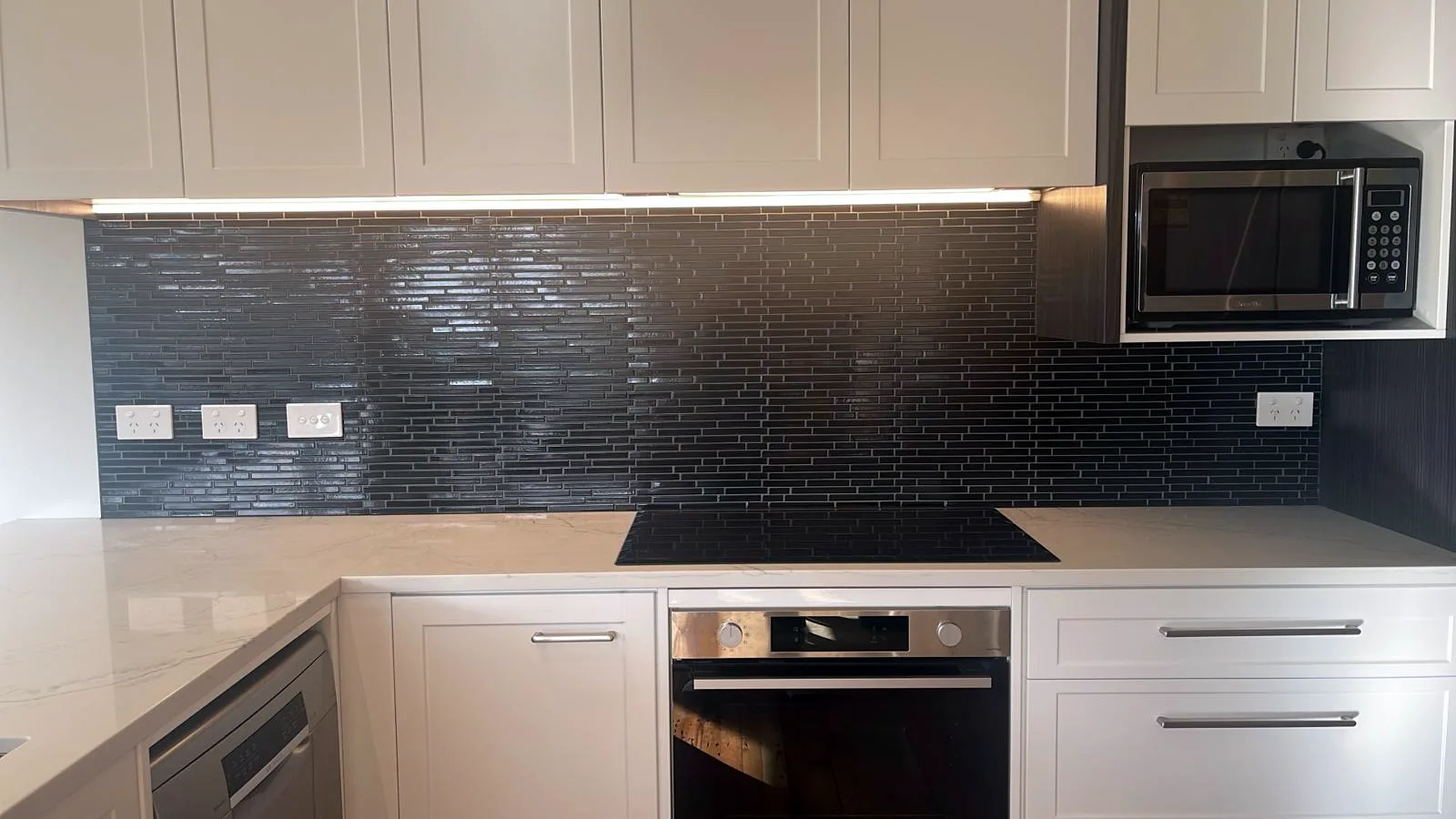 Black glass linear-mosaic kitchen splashback with white shaker cabinetry and stone-look benchtop, Auckland — tiling by Tarara Tiling