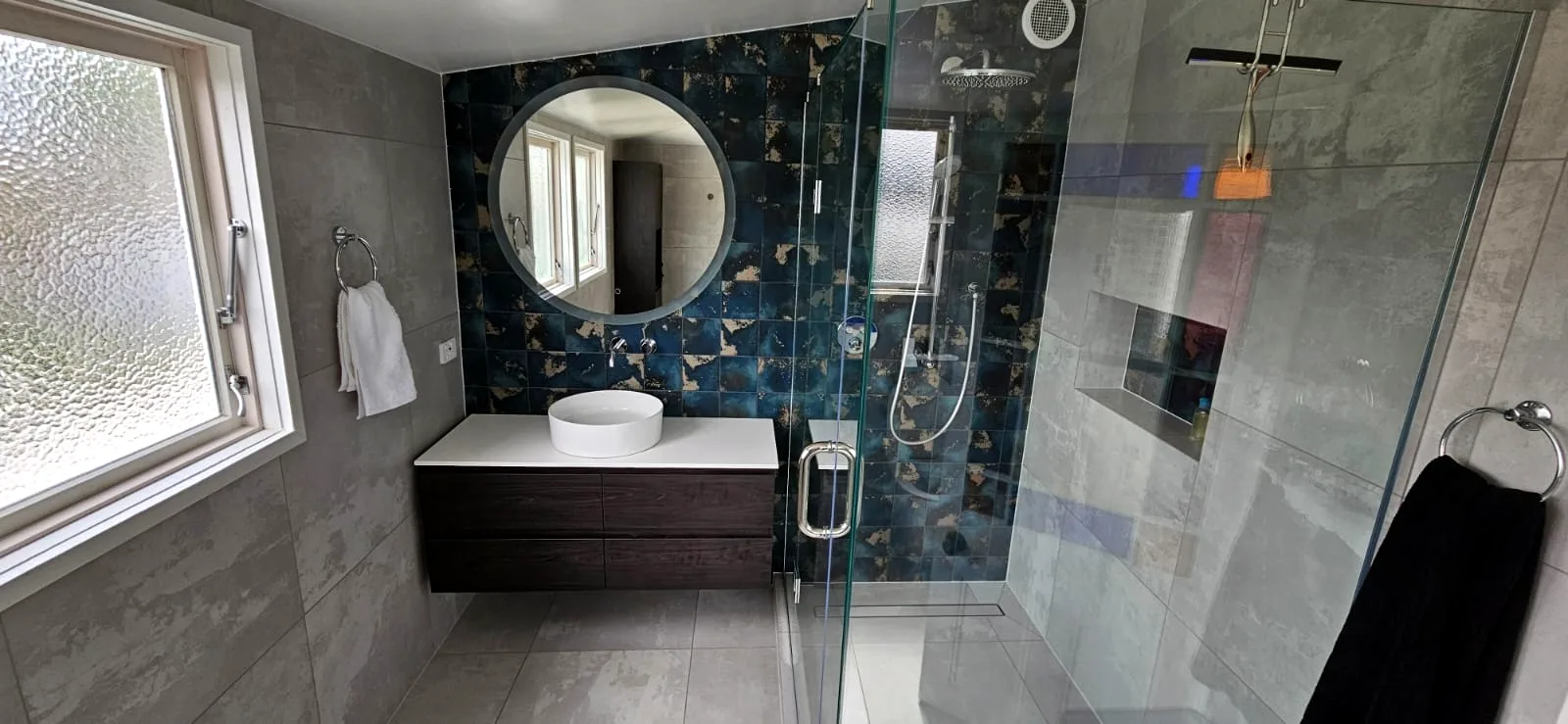 Renovated Auckland bathroom with a glazed blue feature-tile wall, round backlit mirror and walk-in shower — tiling by Tarara Tiling