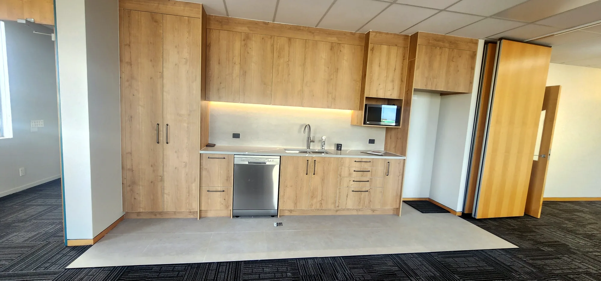 Tiled kitchenette floor with timber cabinetry and integrated appliances in an Auckland commercial fit-out — by Tarara Tiling